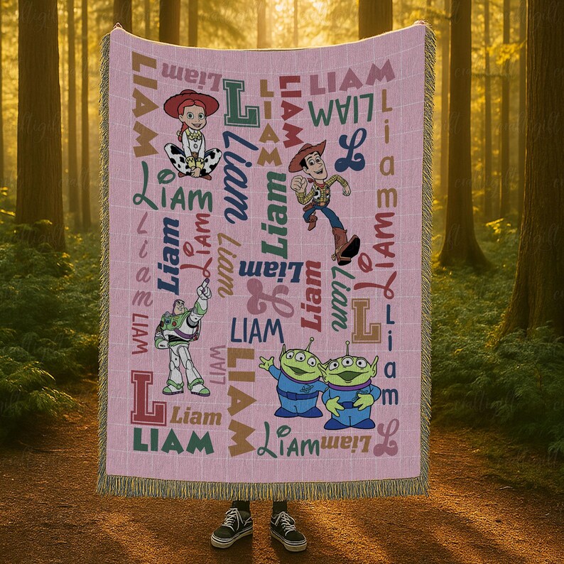 May include: A pink woven throw blanket featuring the name Liam repeated in various fonts and colors alongside illustrations of Jessie, Woody, Buzz Lightyear, and two green aliens from Toy Story. The blanket has a fringed edge on the top and bottom.