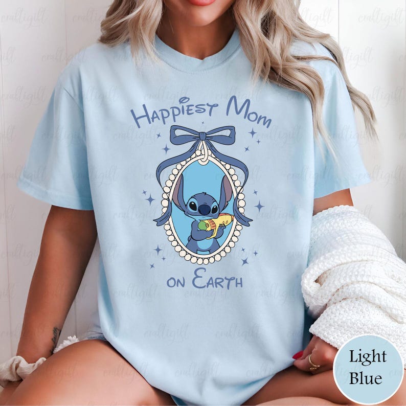 May include: Light blue t-shirt featuring a cartoon image of Stitch holding a snack, inside an oval frame with a bow. The text "Happiest Mom on Earth" is printed above and below the frame. The shirt is a light blue color.