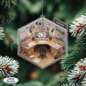May include: Clear acrylic ornament shaped like a miniature artist's studio. The studio features a desk, computer, art supplies, and the word "NAME" on a sign. The ornament is suspended by a silver string against a backdrop of evergreen branches.