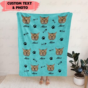 Op de afbeelding: Een turquoise deken met een herhaald patroon van een kattengezicht, pootafdrukken en visgraten. De naam "Alice" staat onder elk kattengezicht afgedrukt. De linkerbovenhoek heeft de tekst "CUSTOM TEXT & PHOTO".