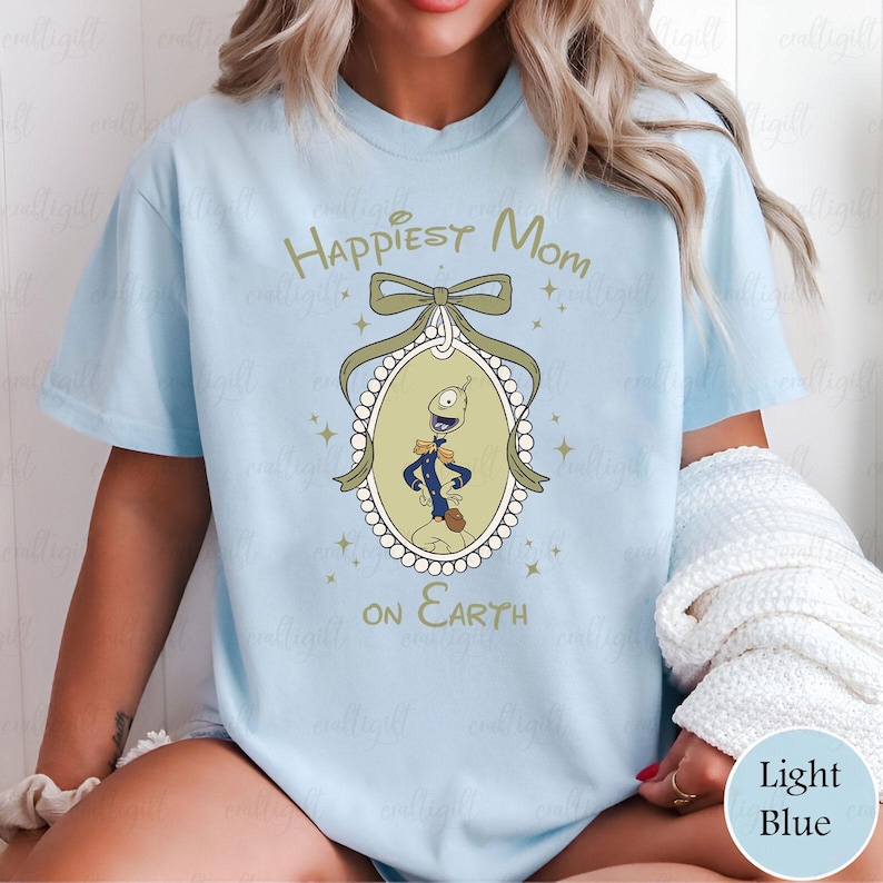 May include: Light blue t-shirt with the text "Happiest Mom on Earth". The shirt features a cartoon character in an oval frame with a bow and star accents. The shirt is a light blue color.
