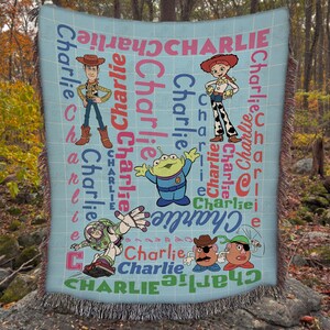 May include: A light blue woven throw blanket featuring Toy Story characters including Woody, Jessie, an alien, Buzz Lightyear, and Mr. and Mrs. Potato Head. The name Charlie is printed repeatedly in various fonts, sizes, and colors across the fabric.