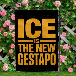 May include: Black garden flag with gold text that reads "ICE IS THE NEW GESTAPO." The flag is hanging on a black metal stand in front of a background of pink roses and green foliage.