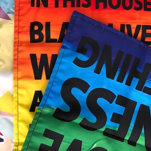 May include: Two fabric flags with rainbow colors and black text. The top flag says "In this house we live with love, kindness, and truth." The bottom flag says "Black Lives Matter."