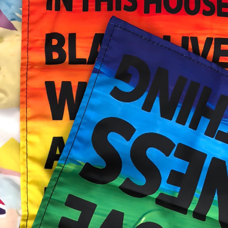 May include: Two fabric flags with rainbow colors and black text. The top flag says "In this house we live with love, kindness, and truth." The bottom flag says "Black Lives Matter."