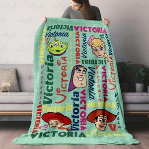 以下が含まれることがあります： 白のグリッドパターンと様々な色やフォントでVictoriaという名前がプリントされたライトグリーンのスローブランケット。ブランケットには、エイリアン、ブロンドの人形、スペースレンジャー、カウガール、カウボーイなどの漫画キャラクターの頭部が描かれています。