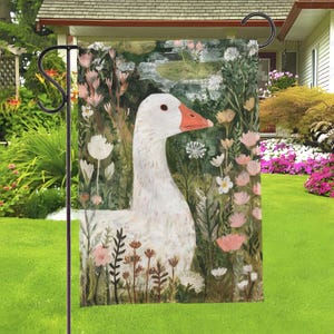 May include: A garden flag featuring a white goose with an orange beak, set against a backdrop of flowers and foliage in shades of green, pink, and white. The flag is displayed on a black metal stand, perfect for outdoor decor.
