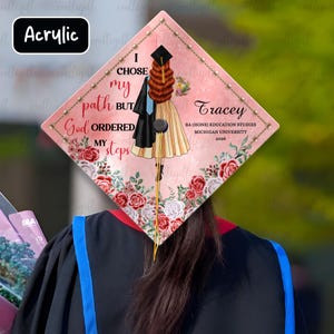 Op de afbeelding: Een roze afstudeerhoed met bloemen- en tekstdetails. De hoed heeft de tekst "I chose my path but God ordered my steps" en de naam "Tracey" met afstudeergegevens. Het woord "Acrylic" staat in de linkerbovenhoek.