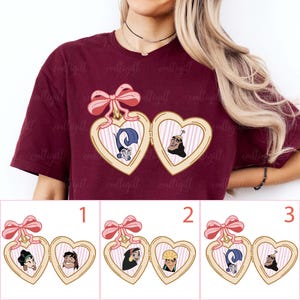 May include: Burgundy t-shirt featuring a heart-shaped locket design with cartoon characters and a pink bow. The locket has a gold frame and a striped background. Below the main design are three additional locket options.