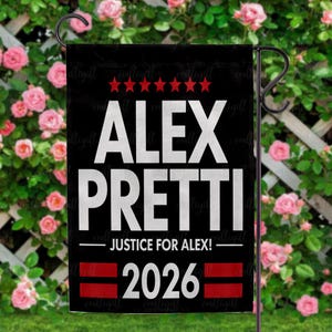 May include: Black garden flag with white text reading "ALEX PRETTI" above "JUSTICE FOR ALEX!" and "2026." The flag has red stars and stripes. The flag is displayed on a black metal stand in front of a rose bush.