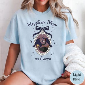May include: Light blue t-shirt with the text "Happiest Mom on Earth" and a cartoon image of a character in an oval frame. The frame is decorated with a bow and stars. The shirt is a light blue color.