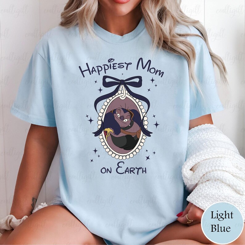 May include: Light blue t-shirt with the text "Happiest Mom on Earth" and a cartoon image of a character in an oval frame. The frame is decorated with a bow and stars. The shirt is a light blue color.