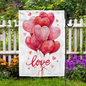May include: A white garden flag featuring a watercolor design of heart-shaped balloons in shades of red and pink. The word "love" is written in a cursive font below the balloons. Small hearts are scattered around the design.