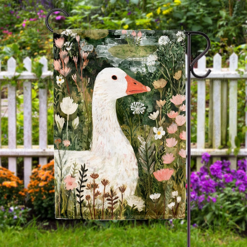 May include: A garden flag featuring a white goose with an orange beak, surrounded by flowers and foliage. The artwork has a watercolor style, with a pond in the background. The flag is displayed on a black metal stand.