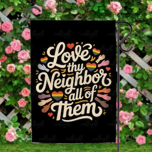 Op de afbeelding: Een zwarte tuinvaandel met de tekst "Love thy Neighbor all of Them" in witte letters. Het ontwerp bevat regenbooghartjes en handen van verschillende huidtinten, met een zwarte metalen standaard en een achtergrond van roze rozen.
