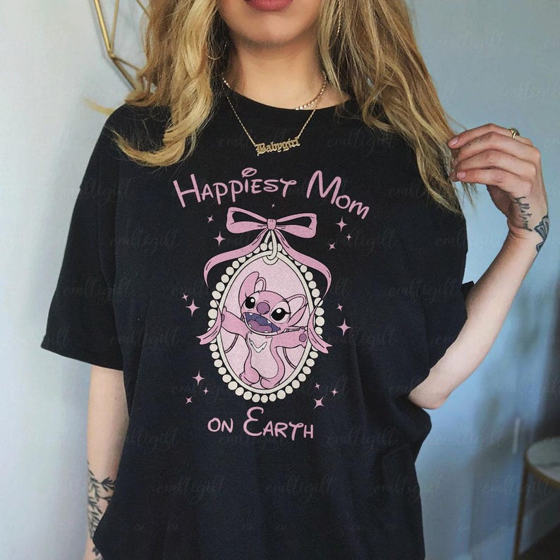 May include: Black t-shirt with a pink graphic of a cartoon character inside a pink oval frame, with the text "Happiest Mom on Earth". The design includes a pink bow and stars.