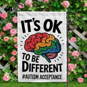May include: White garden flag with the text "IT'S OK TO BE DIFFERENT" and "#AUTISM ACCEPTANCE" in black. A colorful brain graphic is in the center. The flag is hanging on a black metal garden stake, with a background of pink roses.