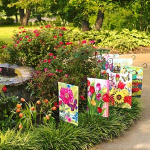 May include: A collection of colorful garden flags with floral and butterfly designs. The flags are displayed on metal poles in a garden setting.