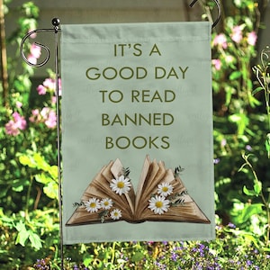 May include: A garden flag with the text "IT'S A GOOD DAY TO READ BANNED BOOKS" in dark green. The flag features an open book illustration with daisies and greenery. The flag is hanging on a black metal stand.