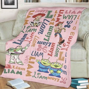 May include: A light pink fleece blanket featuring a white grid pattern, the name Liam in various fonts and colors, and illustrations of Toy Story characters including Woody, Jessie, Buzz Lightyear, and two green aliens scattered across the fabric.