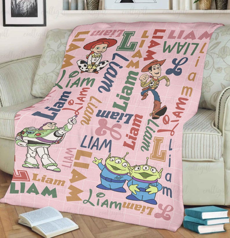 May include: A light pink fleece blanket featuring a white grid pattern, the name Liam in various fonts and colors, and illustrations of Toy Story characters including Woody, Jessie, Buzz Lightyear, and two green aliens scattered across the fabric.