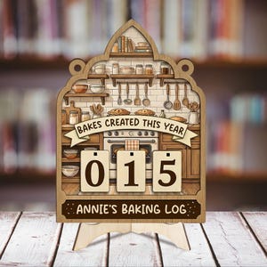 May include: Wooden baking log with a kitchen scene, displaying "Bakes Created This Year" and "015." The sign includes the text "Annie's Baking Log" and is supported by a wooden stand. The background shows a blurred library.