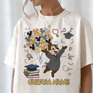 Puede incluir: Camiseta color crema con un personaje de dibujos animados Chipmunk con toga de graduaci&oacute;n, sosteniendo globos de Mickey Mouse. El dise&ntilde;o incluye elementos tem&aacute;ticos de graduaci&oacute;n y el texto "CUSTOM NAME".