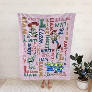 May include: A light pink blanket featuring a repeating pattern of the name Liam in various fonts and colors, alongside illustrations of Jessie, Woody, Buzz Lightyear, and two green aliens from the Toy Story franchise, all on a grid background.
