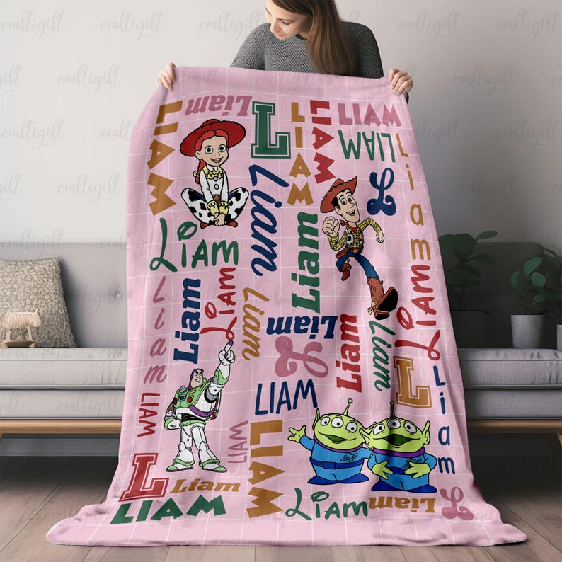 May include: A soft pink fleece blanket featuring a grid pattern and the name Liam printed in various fonts and colors. The blanket displays illustrations of Jessie, Woody, Buzz Lightyear, and two green aliens from the Toy Story film franchise.