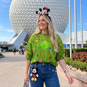 May include: A bright green long-sleeve sweatshirt featuring a repeating floral pattern and a central circular emblem, paired with blue denim jeans. A headband with two small plush character figures sits atop the head, and a plush character keychain hangs from the jeans.
