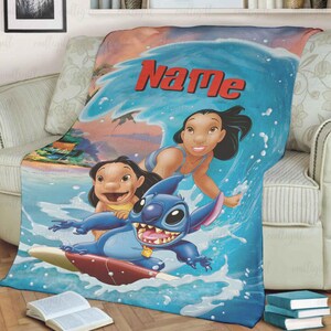 Puede incluir: Una manta azul y blanca con personajes de la pel&iacute;cula animada Lilo & Stitch. La manta representa a Stitch surfeando una ola con Lilo y otro personaje. La palabra "Name" est&aacute; escrita en rojo en la parte superior de la ola.