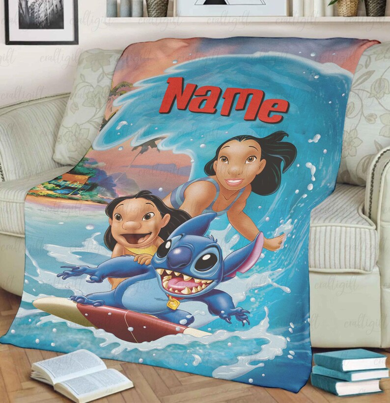 Puede incluir: Una manta azul y blanca con personajes de la pel&iacute;cula animada Lilo & Stitch. La manta representa a Stitch surfeando una ola con Lilo y otro personaje. La palabra "Name" est&aacute; escrita en rojo en la parte superior de la ola.