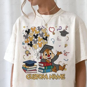 Puede incluir: Camiseta color crema con un dise&ntilde;o tem&aacute;tico de graduaci&oacute;n. El gr&aacute;fico incluye una ardilla de dibujos animados con toga de graduaci&oacute;n, libros, globos y el texto "CUSTOM NAME". El dise&ntilde;o incorpora varios colores y elementos de celebraci&oacute;n.