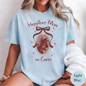 May include: Light blue t-shirt with the text "Happiest Mom on Earth" and a cartoon character illustration in a decorative oval frame. The illustration features a red and brown creature. The shirt has short sleeves.