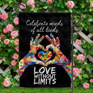 May include: Black garden flag with the text "Celebrate minds of all kinds" and "Love Without Limits." The flag features a colourful heart formed by two hands with a patchwork design. The background is a lush green garden with pink roses.