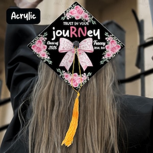 May include: A black graduation cap decorated with pink floral accents and the words "TRUST IN YOUR JOURNEY". A pink bow with medical-themed illustrations is centered. The tassel is yellow. The text "Class of 2026" and a name are also visible.
