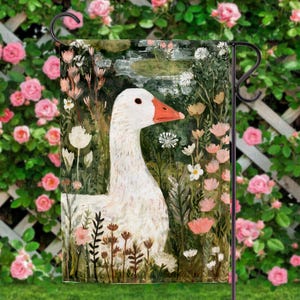 May include: A garden flag featuring a white goose with an orange beak, set against a backdrop of lush greenery and pink and white flowers. The artwork has a painterly style, with visible brushstrokes. The flag is displayed on a black metal stand, perfect for outdoor decor.