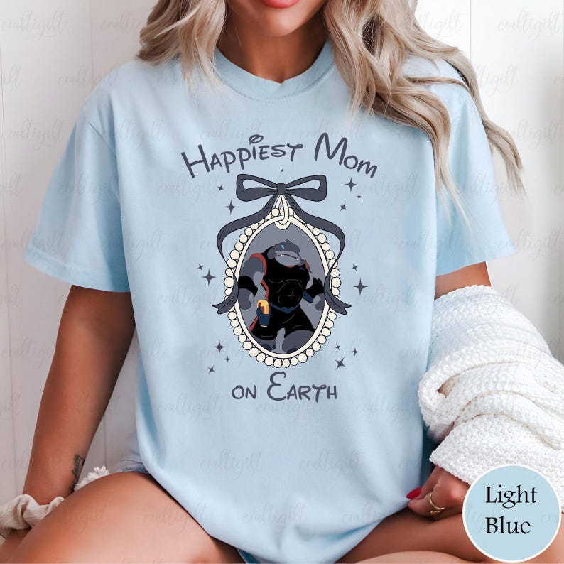 May include: Light blue t-shirt with the text "Happiest Mom on Earth". The shirt features a cartoon illustration of a character in a black and red outfit inside an oval frame with a bow and star accents. The shirt is a light blue color.