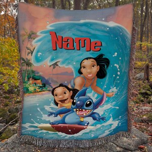 Puede incluir: Una manta tejida con una vibrante ilustraci&oacute;n de personajes surfeando una ola. El dise&ntilde;o incluye una ola azul, personajes y la palabra "Name" en rojo. La manta tiene un borde con flecos y una colorida escena tropical.