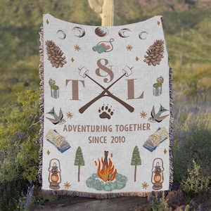 May include: A light grey woven throw blanket with a camping theme. The blanket features pine cones, binoculars, a paw print, and the text "Adventuring Together Since 2010". The blanket also has a campfire, trees, and lanterns.