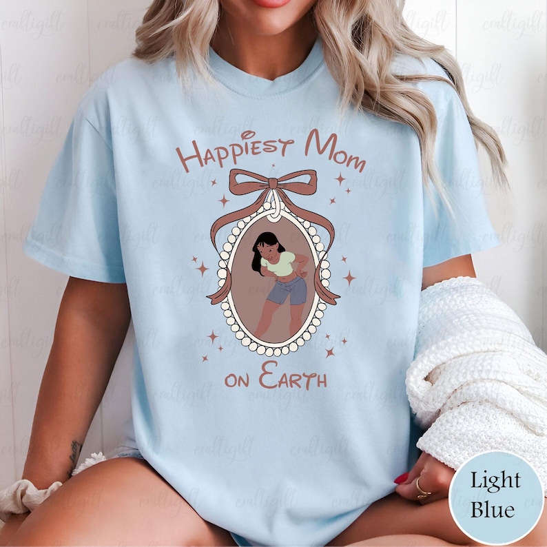 May include: Light blue t-shirt with the text "Happiest Mom on Earth". The shirt features a framed illustration of a person in a light green top and blue shorts. The frame is brown with a bow at the top and is surrounded by small star shapes.