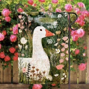 May include: A garden flag featuring a white goose with an orange beak, surrounded by pink and white flowers and greenery. The artwork has a painterly style, with a wooden fence and rose bushes in the background. The flag is designed for outdoor use.