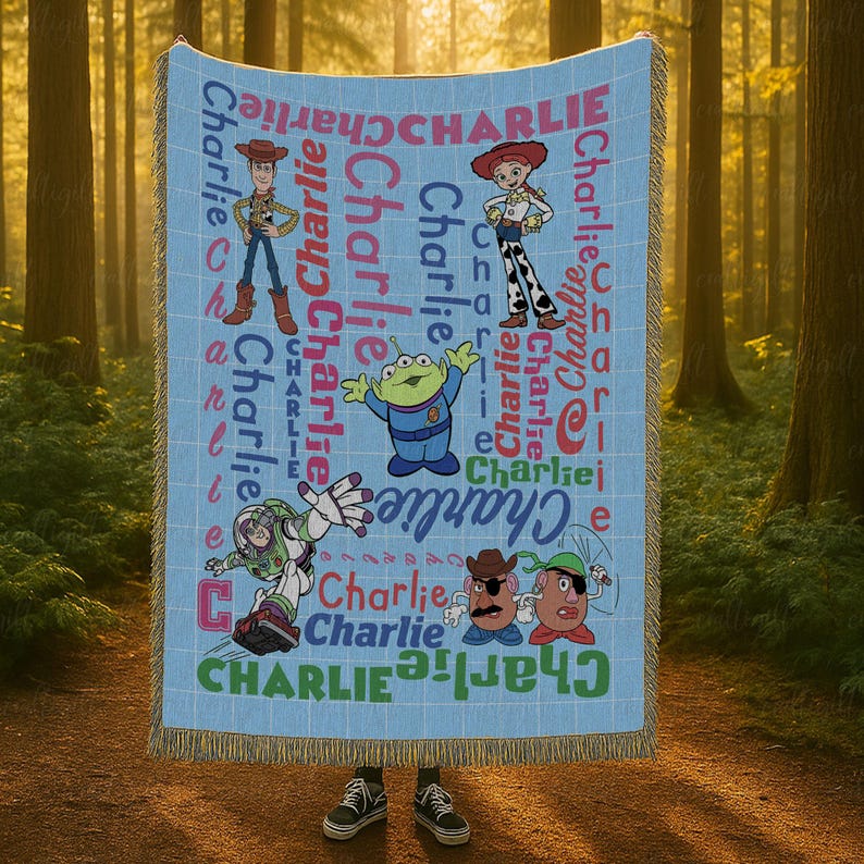 May include: A light blue woven throw blanket featuring a grid pattern and the name Charlie repeated in various colors and fonts. The blanket displays cartoon characters including a cowboy, a cowgirl, a green alien, a space ranger, and two potatoes.