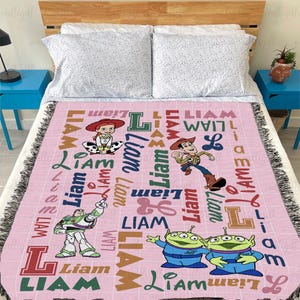May include: A light pink woven throw blanket featuring the name Liam repeated in various fonts and colors, alongside printed illustrations of Jessie, Woody, Buzz Lightyear, and two green aliens from the Toy Story film franchise.