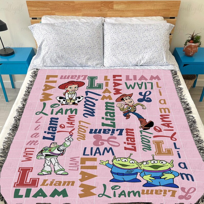 May include: A light pink woven throw blanket featuring the name Liam repeated in various fonts and colors, alongside printed illustrations of Jessie, Woody, Buzz Lightyear, and two green aliens from the Toy Story film franchise.