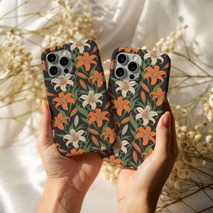 May include: Two black phone cases with a floral pattern featuring orange and white lilies and green leaves. The cases are held up, showcasing the design on a dark background.