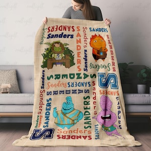 May include: A beige throw blanket featuring a grid pattern and the repeated name Sanders in various colors and fonts. The blanket displays four cartoon characters: a green creature at a desk, an orange flame-like figure, a blue character, and a pink blob.