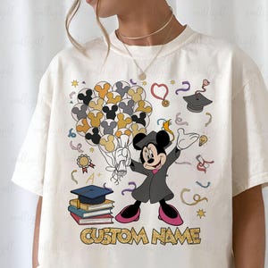 Puede incluir: Camiseta color crema con un dise&ntilde;o de Mickey Mouse con tem&aacute;tica de graduaci&oacute;n. Mickey lleva una toga de graduaci&oacute;n y sostiene un diploma y globos. El dise&ntilde;o incluye birretes, libros y el texto "CUSTOM NAME".
