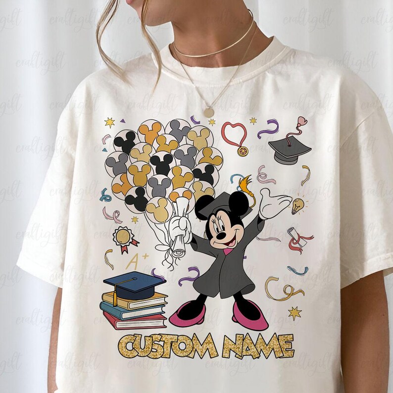Puede incluir: Camiseta color crema con un dise&ntilde;o de Mickey Mouse con tem&aacute;tica de graduaci&oacute;n. Mickey lleva una toga de graduaci&oacute;n y sostiene un diploma y globos. El dise&ntilde;o incluye birretes, libros y el texto "CUSTOM NAME".