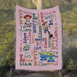 May include: A woven throw blanket in a light pink hue featuring a repeating pattern of the name Liam in various fonts and colors, alongside illustrations of Jessie, Woody, Buzz Lightyear, and two green aliens from the Toy Story film franchise.
